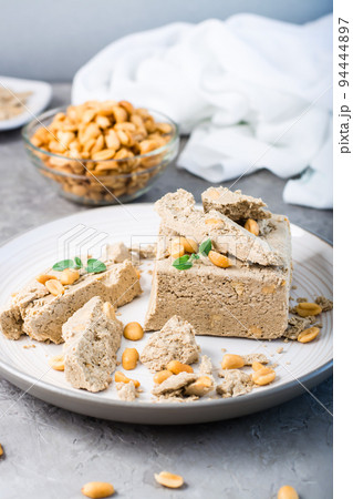 Pieces of sunflower and peanut halva and mint leaves on a plate and a bowl of nuts on the table. Caloric oriental delicacy. Vertical view. Close-up 94444897