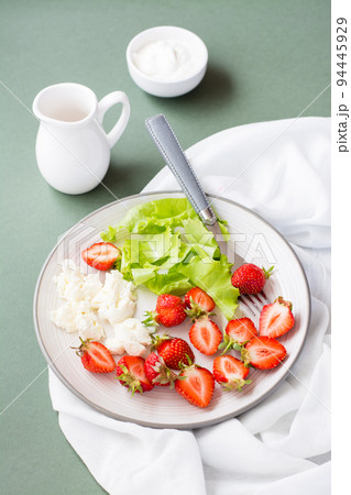 Fresh whole and sliced strawberries, cottage cheese and lettuce on a plate and sour cream in a bowl on a green background. Summer vitamin snack. Vertical view 94445929