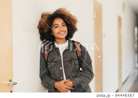 Young student with backpack smiling in high school leaning a wall	 94446367