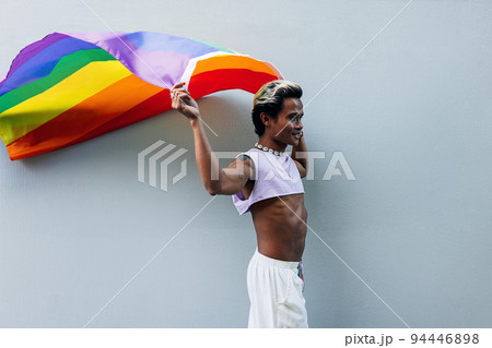 Stylish guy waving LGBT flag while walking at grey wall outdoors	 94446898