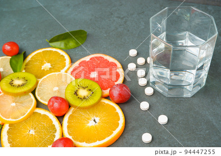 Supplements or healthy natural food? Scattered white pills vitamins, Sliced citrus fruits, orange, lemon, kiwi, grapes, glass with water on a stone gray background Supplements or healthy natural food? Scattered white pills vitamins, Sliced citrus fruits, orange, lemon, kiwi, grapes, glass with water on a stone gray background 94447255