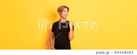 Portrait of pleased thoughtful asian guy, looking and pointing finger left at logo, standing yellow background Portrait of pleased thoughtful asian guy, looking and pointing finger left at logo, standing yellow background 94448085