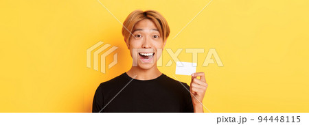 Close-up of excited surprised handsome asian guy, smiling amazed and showing credit card, standing yellow background 94448115