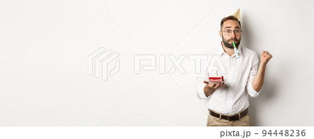 Holidays and celebration. Cheerful man enjoying birthday, blowing party whistle and holding bday cake, white background Holidays and celebration. Cheerful man enjoying birthday, blowing party whistle and holding bday cake, white background 94448236