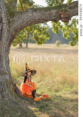 A girl in guise of witch is laughing merrily sitting under huge tree trunk on edge of forest with carved frightening pumpkins. In anticipation of Halloween holiday A girl in guise of witch is laughing merrily sitting under huge tree trunk on edge of forest with carved frightening pumpkins. In anticipation of Halloween holiday 94448874