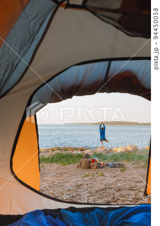 view from a tent on the shore of a lake or sea, a woman collects firewood or does yoga and meditation. camping in nature 94450058
