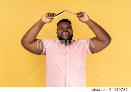 Portrait of joyful delighted bearded man wearing pink shirt holding opened book on head, looking up with happy positive expression. Indoor studio shot isolated on yellow background. Portrait of joyful delighted bearded man wearing pink shirt holding opened book on head, looking up with happy positive expression. Indoor studio shot isolated on yellow background. 94450786