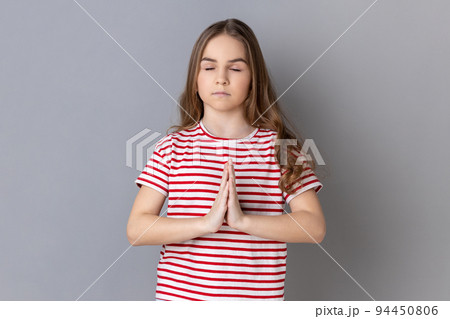 Portrait of little girl wearing striped T-shirt keeping hands namaste gesture, meditating, yoga exercise breath technique reduce stress. Indoor studio shot isolated on gray background. 94450806