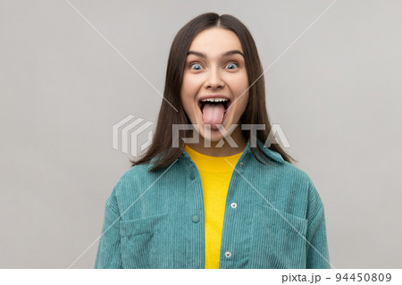 Dark haired woman sticking out tongue and looking at camera, teasing with naughty expression, disobedient behavior, wearing casual style jacket. Indoor studio shot isolated on gray background. Dark haired woman sticking out tongue and looking at camera, teasing with naughty expression, disobedient behavior, wearing casual style jacket. Indoor studio shot isolated on gray background. 94450809