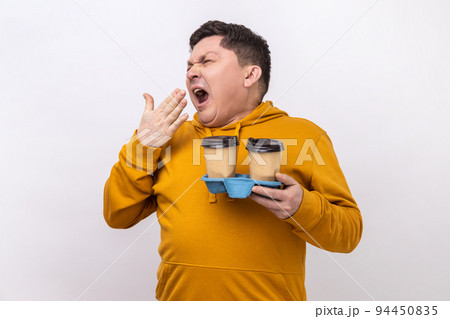 Portrait of tired exhausted man holding coffee cups and yawning, feeling bored after stressful working day, wearing urban style hoodie. Indoor studio shot isolated on white background. Portrait of tired exhausted man holding coffee cups and yawning, feeling bored after stressful working day, wearing urban style hoodie. Indoor studio shot isolated on white background. 94450835