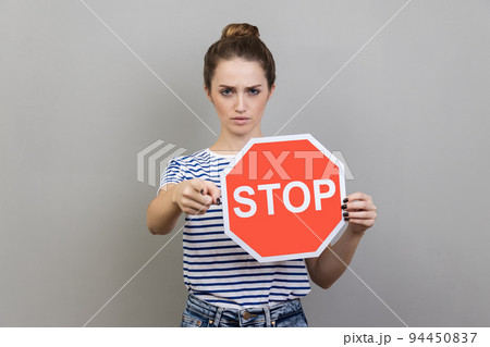 Portrait of serious bossy woman with bun hairstyle wearing striped T-shirt holding red stop sign pointing at camera, has strict expression. Indoor studio shot isolated on gray background. Portrait of serious bossy woman with bun hairstyle wearing striped T-shirt holding red stop sign pointing at camera, has strict expression. Indoor studio shot isolated on gray background. 94450837