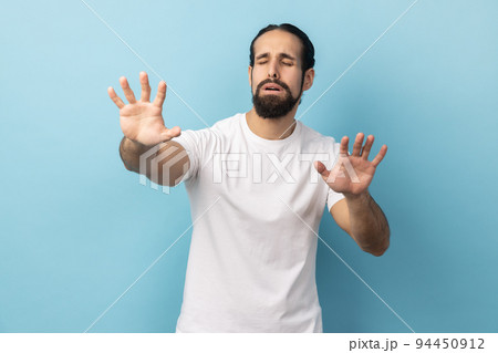 Portrait of disoriented blind man with beard wearing white T-shirt standing with stretched arms, searching for way, vision problems. Indoor studio shot isolated on blue background. Portrait of disoriented blind man with beard wearing white T-shirt standing with stretched arms, searching for way, vision problems. Indoor studio shot isolated on blue background. 94450912