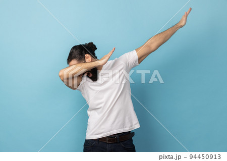 Portrait of anonymous unknown man with beard wearing white T-shirt standing in dab dance pose, internet meme, celebrating success. Indoor studio shot isolated on blue background. 94450913