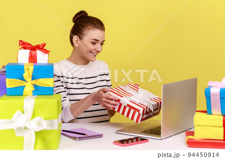 Joyful woman holding gift box and looking at laptop screen, giving birthday present while having online celebration by video call. Indoor studio studio shot isolated on yellow background. 94450914