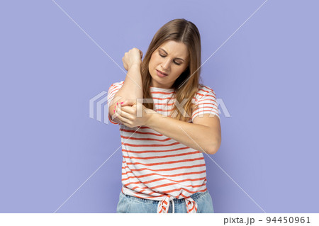 Unhealthy sick blond woman wearing striped T-shirt having trauma after car accident, feels pain in elbow, trapped nerves, rheumatoid arthritis. Indoor studio shot isolated on purple background. Unhealthy sick blond woman wearing striped T-shirt having trauma after car accident, feels pain in elbow, trapped nerves, rheumatoid arthritis. Indoor studio shot isolated on purple background. 94450961