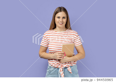 Portrait of cheerful pretty blond woman student wearing striped T-shirt holding book or paper organizer, looking at camera with smile. Indoor studio shot isolated on purple background. 94450965