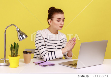 Woman in striped shirt holding hands in praying gesture and talking on video call on laptop, asking heartily, pleading with desperate face. Indoor studio studio shot isolated on yellow background. Woman in striped shirt holding hands in praying gesture and talking on video call on laptop, asking heartily, pleading with desperate face. Indoor studio studio shot isolated on yellow background. 94450974