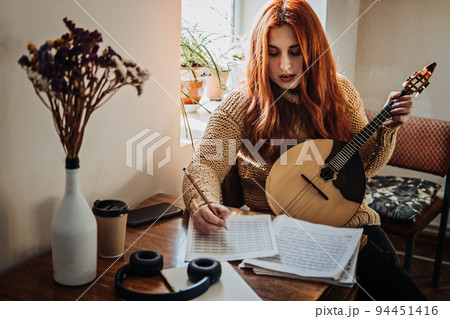 Redhead woman playing acoustic Domra Folk Musical Instrument and looking into note. Young woman taking music lessons at home 94451416