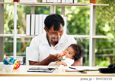 Asian Young father in white t shrit working from home and holding his baby boy with milk bottle Asian Young father in white t shrit working from home and holding his baby boy with milk bottle 94458350