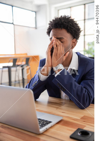 Portrait of young tired office worker at table rubbing nose Portrait of young tired office worker at table rubbing nose 94470396