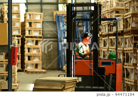 Warehouse worker in uniform loading cardboard boxes with forklift in warehouse full of shelves with goods Warehouse worker in uniform loading cardboard boxes with forklift in warehouse full of shelves with goods 94471104