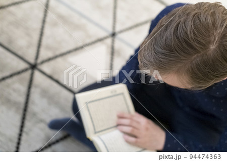 Young muslim man sitting on the floor while reading holy book Quran before iftar dinner during a ramadan feast at home 94474363