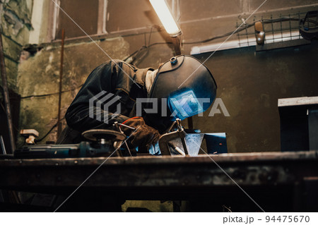 Worker in a welder mask works in a workshop for welding iron. Man makes iron products. Guy works with a welding machine using a mask to protect his eyes from dangerous rays Worker in a welder mask works in a workshop for welding iron. Man makes iron products. Guy works with a welding machine using a mask to protect his eyes from dangerous rays 94475670