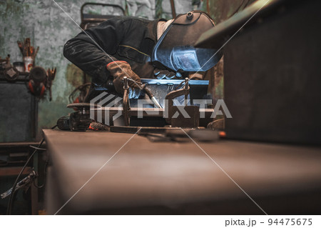 Worker in a welder mask works in a workshop for welding iron. Man makes iron products. Guy works with a welding machine using a mask to protect his eyes from dangerous rays 94475675