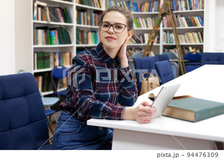 Girl in glasses working with tablet sitting in library. Student prefers to work with a tablet than books. Girl in glasses working with tablet sitting in library. Student prefers to work with a tablet than books. 94476309