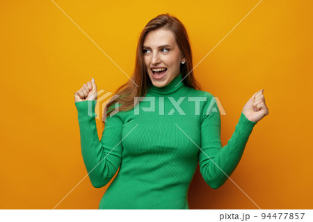 Portrait of an excited young woman celebrating success with raised arms against yellow background 94477857
