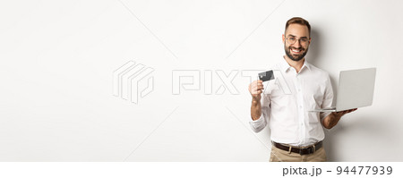 Online shopping. Handsome man showing credit card and using laptop to order in internet, standing over white background Online shopping. Handsome man showing credit card and using laptop to order in internet, standing over white background 94477939