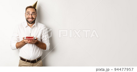 Holidays and celebration. Happy man having birthday party, making wish on b-day cake and smiling, standing against white background Holidays and celebration. Happy man having birthday party, making wish on b-day cake and smiling, standing against white background 94477957