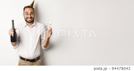 Celebration and holidays. Happy birthday guy enjoying b-day party, wearing funny cone hat and drinking champagne, white background Celebration and holidays. Happy birthday guy enjoying b-day party, wearing funny cone hat and drinking champagne, white background 94478042