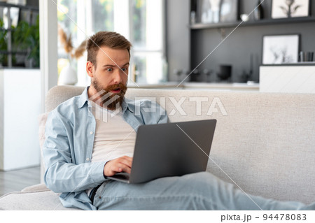 Young bearded man sitting on sofa at home using laptop computer 94478083