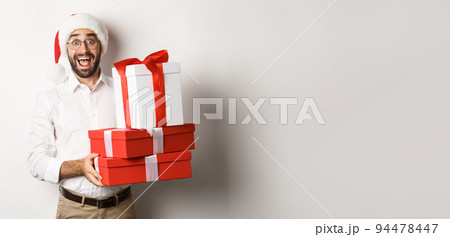 Winter holidays and celebration. Happy guy bring christmas presents, holding gifts and wearing Santa hat, standing over white background Winter holidays and celebration. Happy guy bring christmas presents, holding gifts and wearing Santa hat, standing over white background 94478447