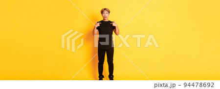 Full-length of surprised attractive korean blond guy in black outfit pointing at himself impressed, yellow background Full-length of surprised attractive korean blond guy in black outfit pointing at himself impressed, yellow background 94478692