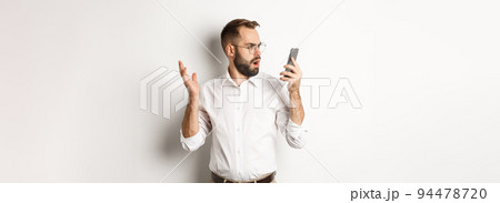 Man looking confused at mobile phone after conversation, standing puzzled over white background Man looking confused at mobile phone after conversation, standing puzzled over white background 94478720