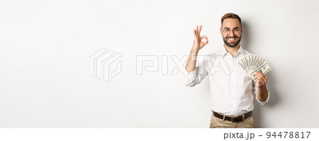 Satisfied young businessman showing money, make okay sign, earning cash, standing over white background Satisfied young businessman showing money, make okay sign, earning cash, standing over white background 94478817