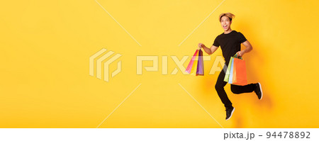 Full-length of happy attractive asian man jumping from happiness and carry shopping bags, standing yellow background Full-length of happy attractive asian man jumping from happiness and carry shopping bags, standing yellow background 94478892