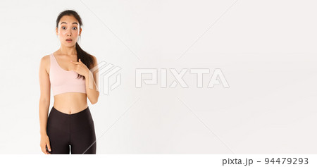 Sport, wellbeing and active lifestyle concept. Surprised asian girl in fitness clothing looking wondered and pointing at herself confused, being chosen or picked, white background 94479293