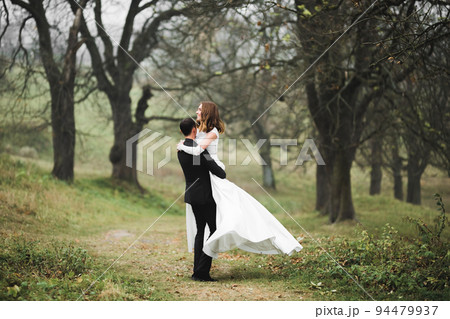 Romantic, fairytale, happy newlywed couple hugging and kissing in a park, trees in background Romantic, fairytale, happy newlywed couple hugging and kissing in a park, trees in background 94479937
