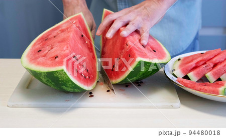Man in home kitchen while cutting a ripe red watermelon in slices. 94480018