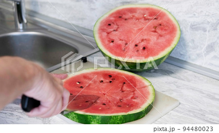 Man in home kitchen while cutting a ripe red watermelon in slices. 94480023