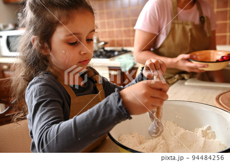 Close-up portrait of an adorable Caucasian little girl mixing dry ingredients with white flour in an enamel bowl, kneading dough in home kitchen, helping her mother in cooking festive tasty cherry pie Close-up portrait of an adorable Caucasian little girl mixing dry ingredients with white flour in an enamel bowl, kneading dough in home kitchen, helping her mother in cooking festive tasty cherry pie 94484256