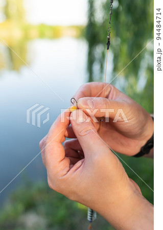 The concept of bait for fish. Close-up of a fisherman's male hand stringing bait on a fishing rod against a blue lake. 94484714