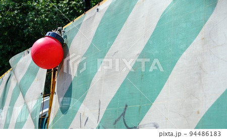 Fence enclosing the construction site for safety reasons, made of thick dense fabric with green stripes and signal red round lights. Fence enclosing the construction site for safety reasons, made of thick dense fabric with green stripes and signal red round lights. 94486183