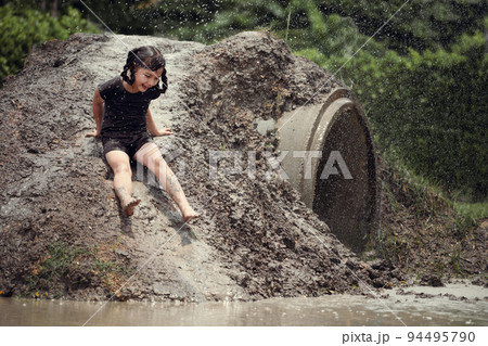 Cheerful child girl playing on heap of wet soil during raining in rainy season. Cheerful child girl playing on heap of wet soil during raining in rainy season. 94495790