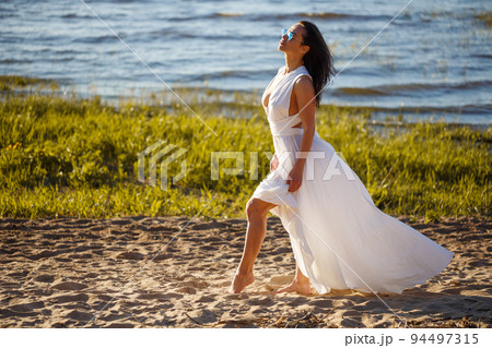 Beautiful woman in white dress posing on the shore Beautiful woman in white dress posing on the shore 94497315