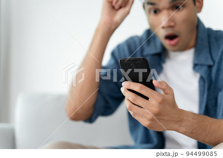 Portrait of excited asian man looking at mobile phone, man receiving good news using smartphone at home. 94499085