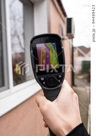 A male hand holds a thermal imager at the window of a house. Search for heat loss in private houses. 94499423